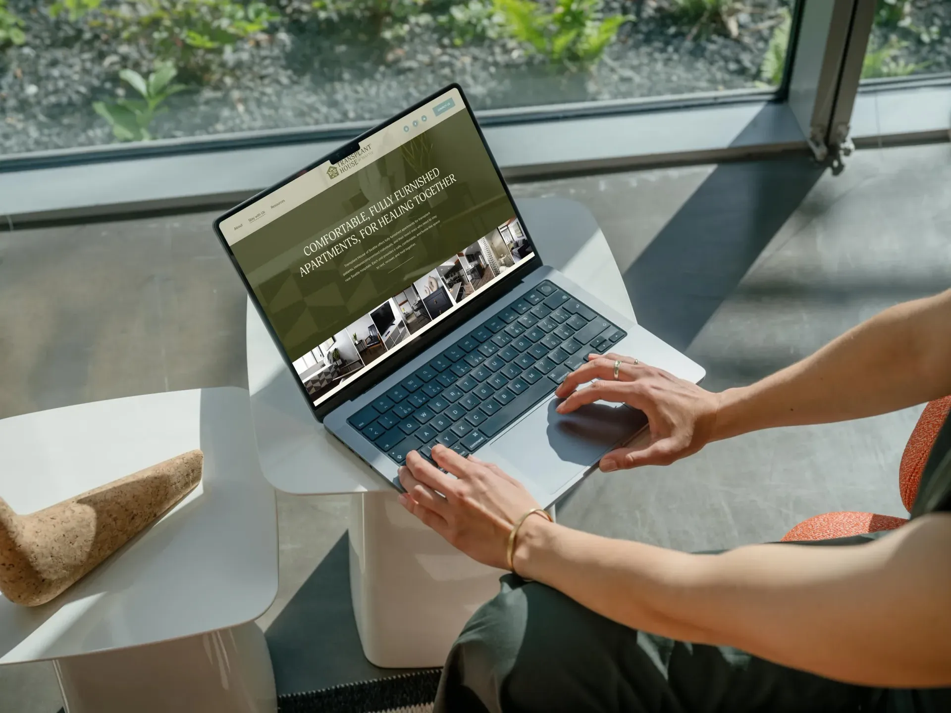 Person using a laptop, typing at a table near a window. Website visible on screen.