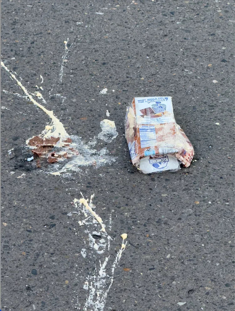 Damaged carton of chocolate milk on asphalt with white liquid splashes.