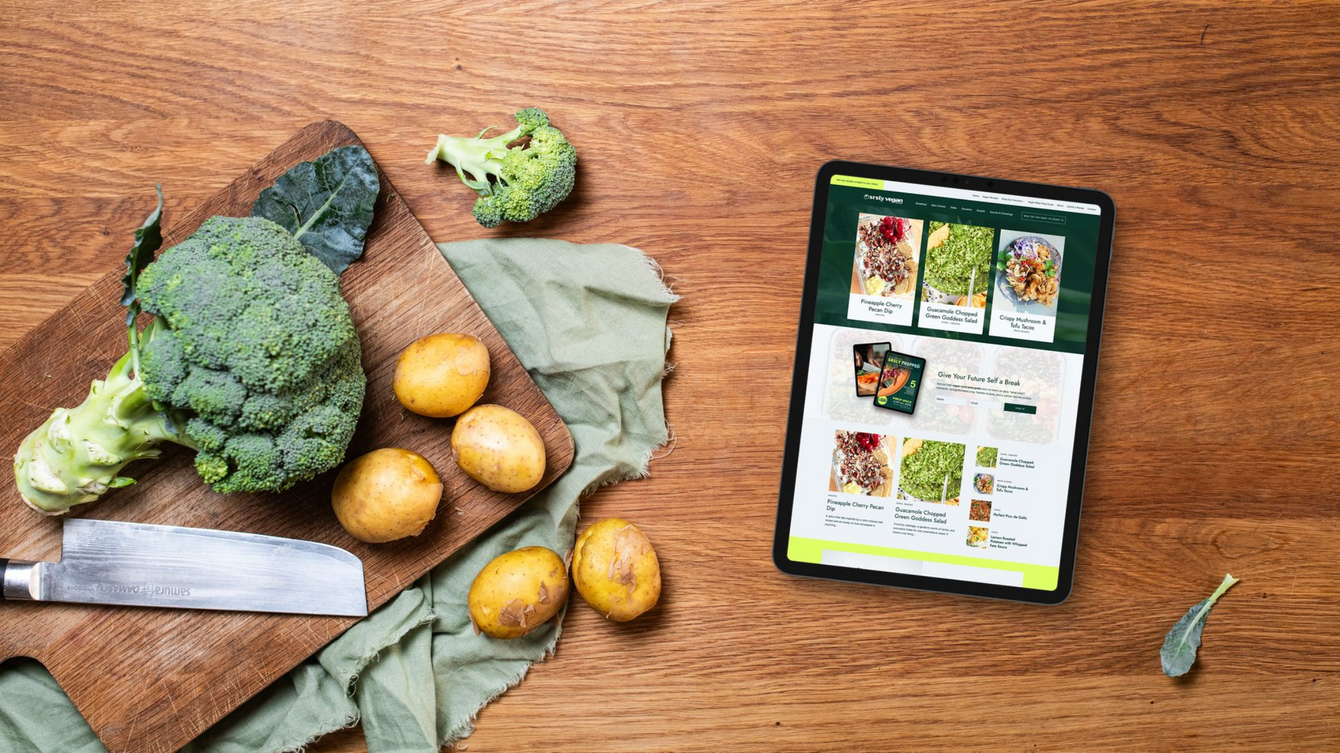 Broccoli, potatoes, and knife on cutting board beside tablet with food images, on a wooden surface.