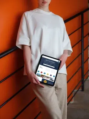 Person holding a tablet displaying a website, standing by an orange wall and black railing.