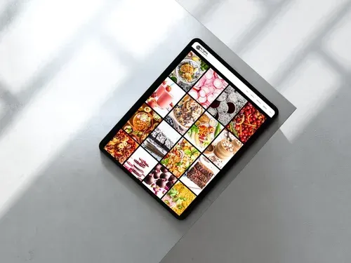 Tablet displaying a grid of food photos on a white surface, lit by shadows.