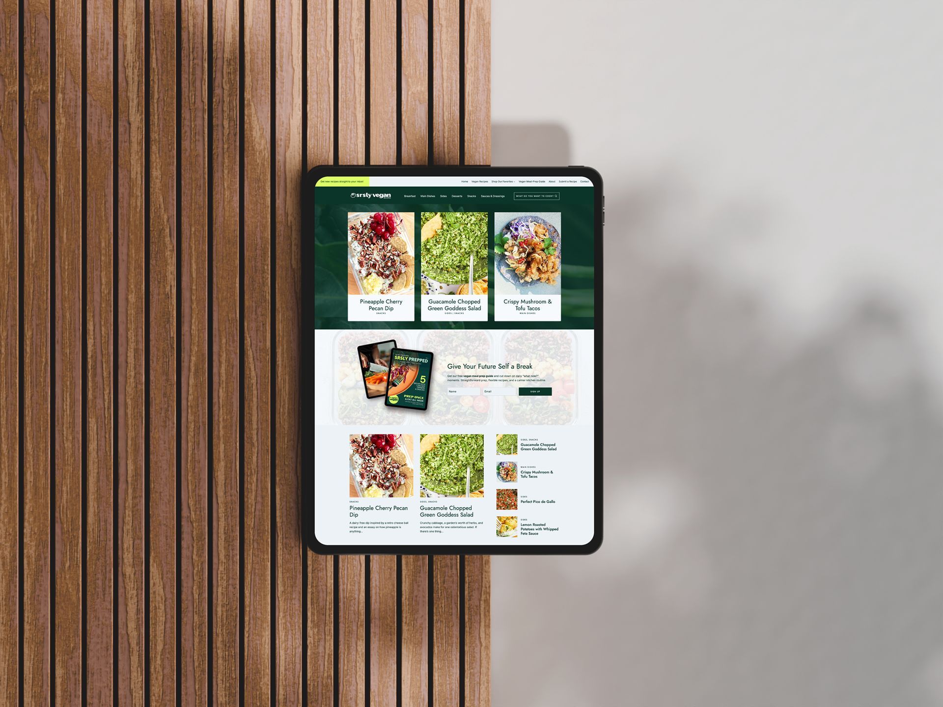Tablet displaying a website with food photos, mounted on a wooden panel next to a white wall.