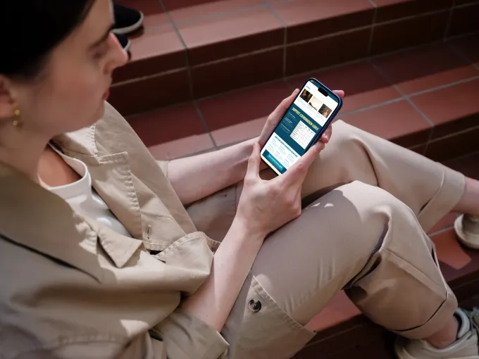 Woman seated on steps, looking at a phone displaying a screen with lists and icons. Beige clothing, natural lighting.