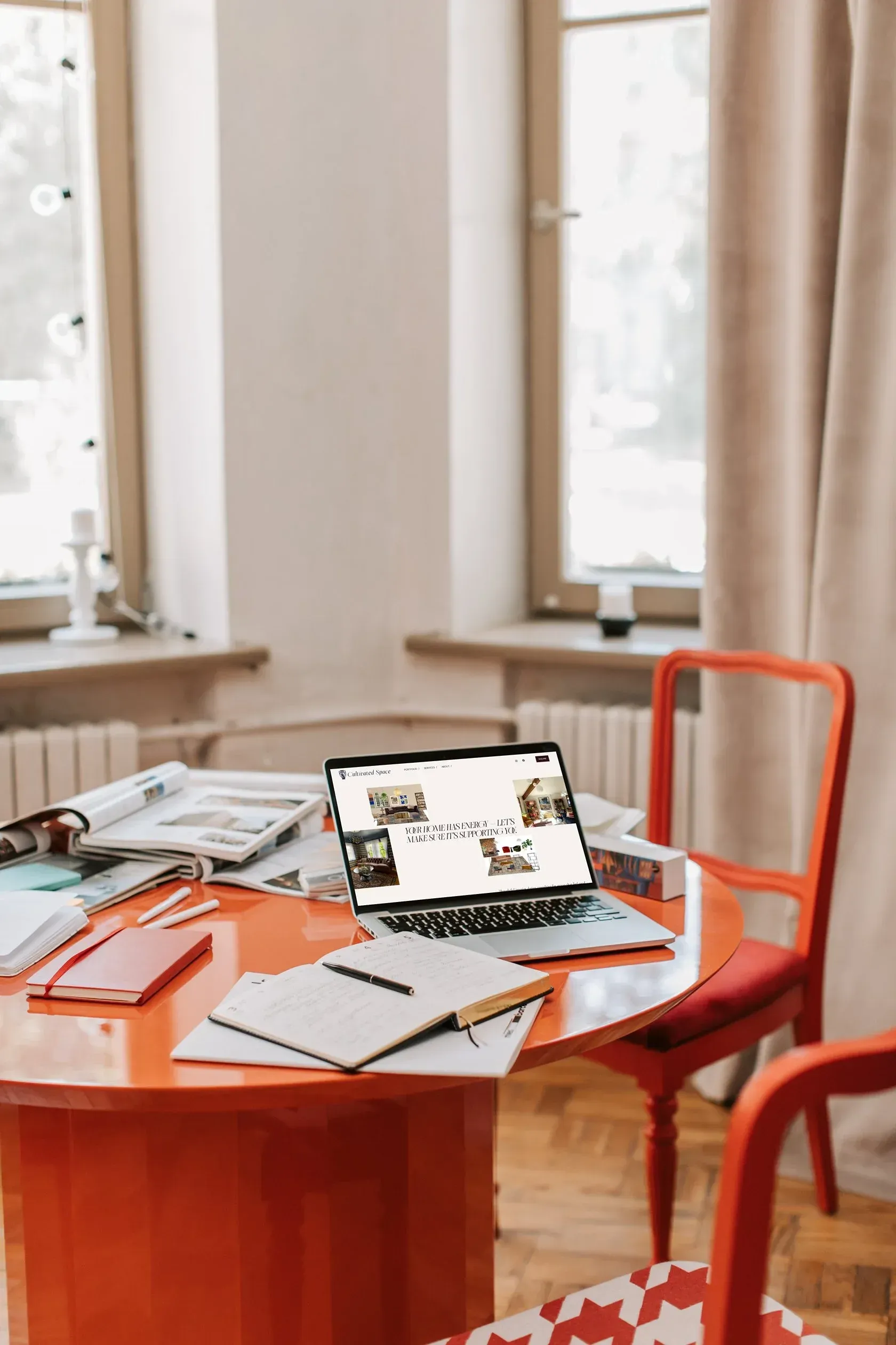 Laptop on a bright orange table with notebooks, magazines, and a red chair by a window.