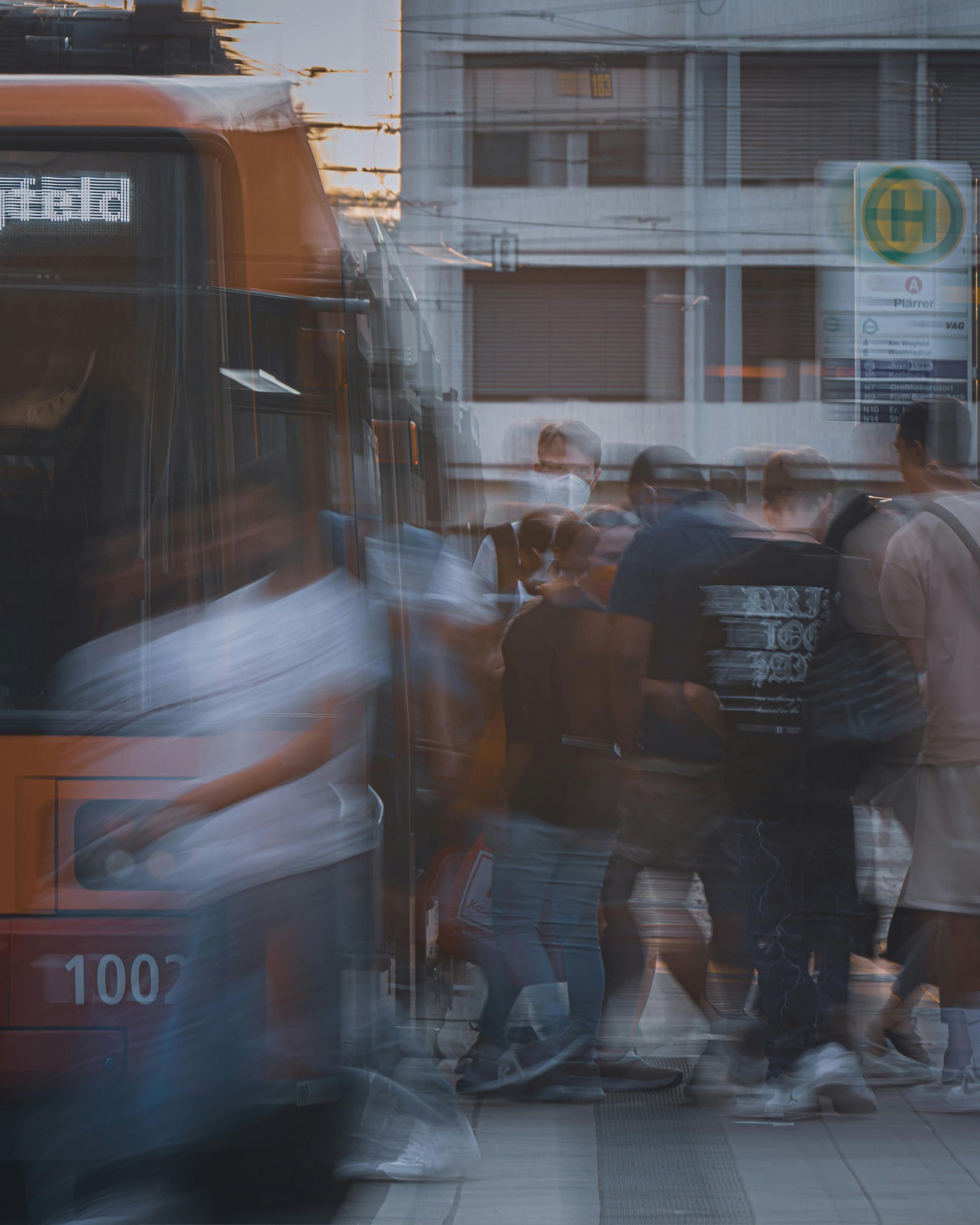 Bus at a busy stop; people blurred by motion, entering/exiting. White, orange, and neutral tones.