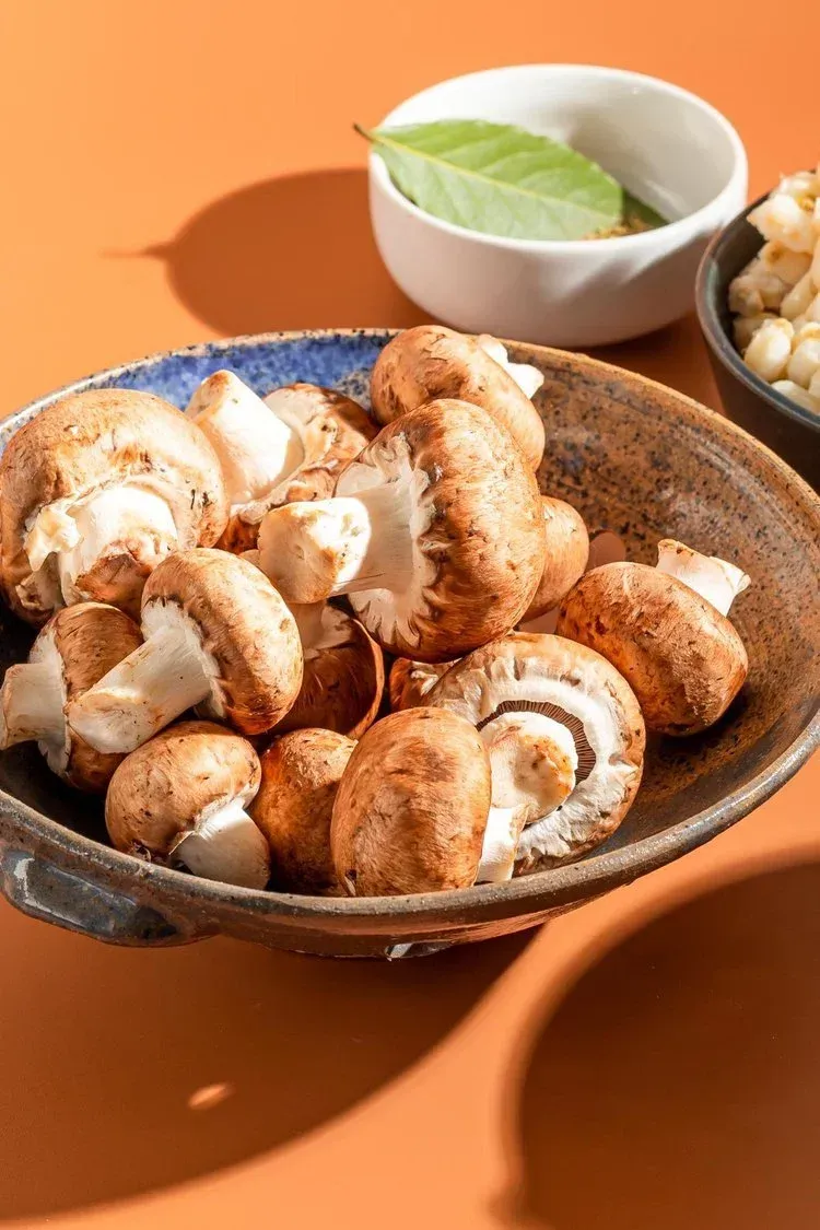 A rustic bowl filled with fresh brown mushrooms sits on an orange surface, with a bay leaf in a small bowl nearby.