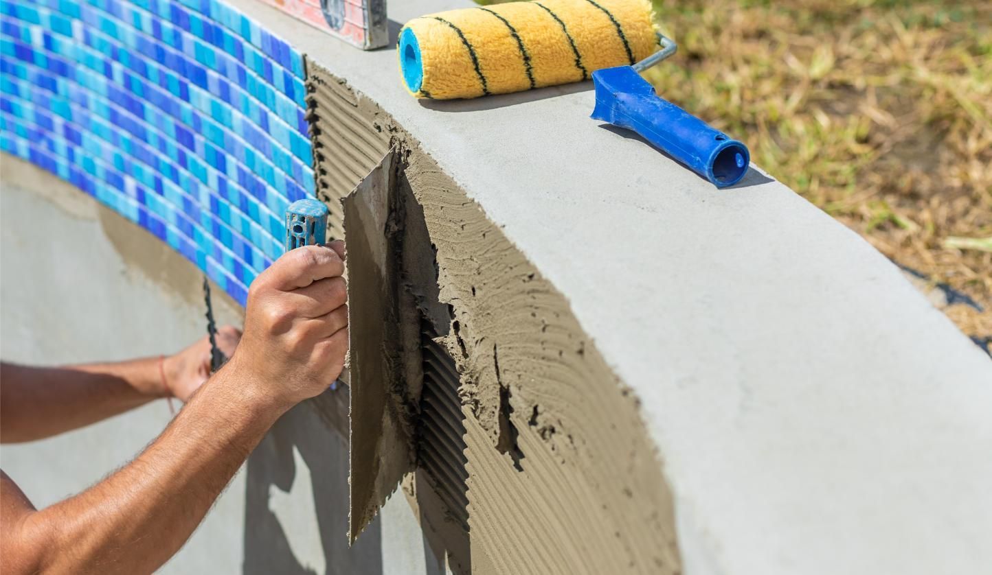 Person tiling a pool's edge with blue mosaic tiles. Mortar is applied to the surface.