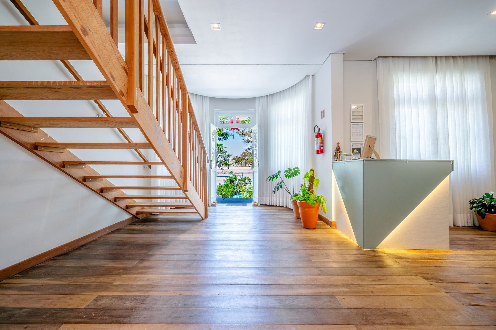 Escadaria de madeira e área de recepção com balcão azul claro e porta aberta com vista para o exterior.