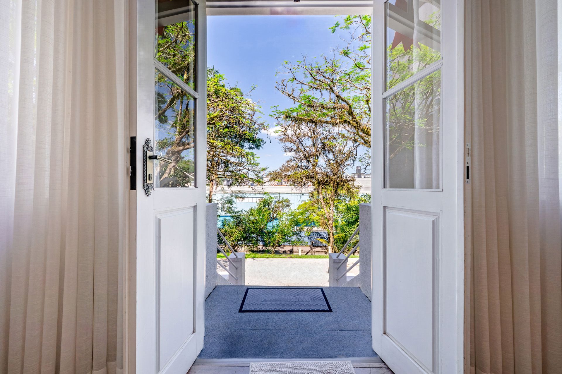 Ao abrir as portas duplas brancas, revela-se uma paisagem ensolarada ao ar livre: árvores, céu azul e um vislumbre da água.