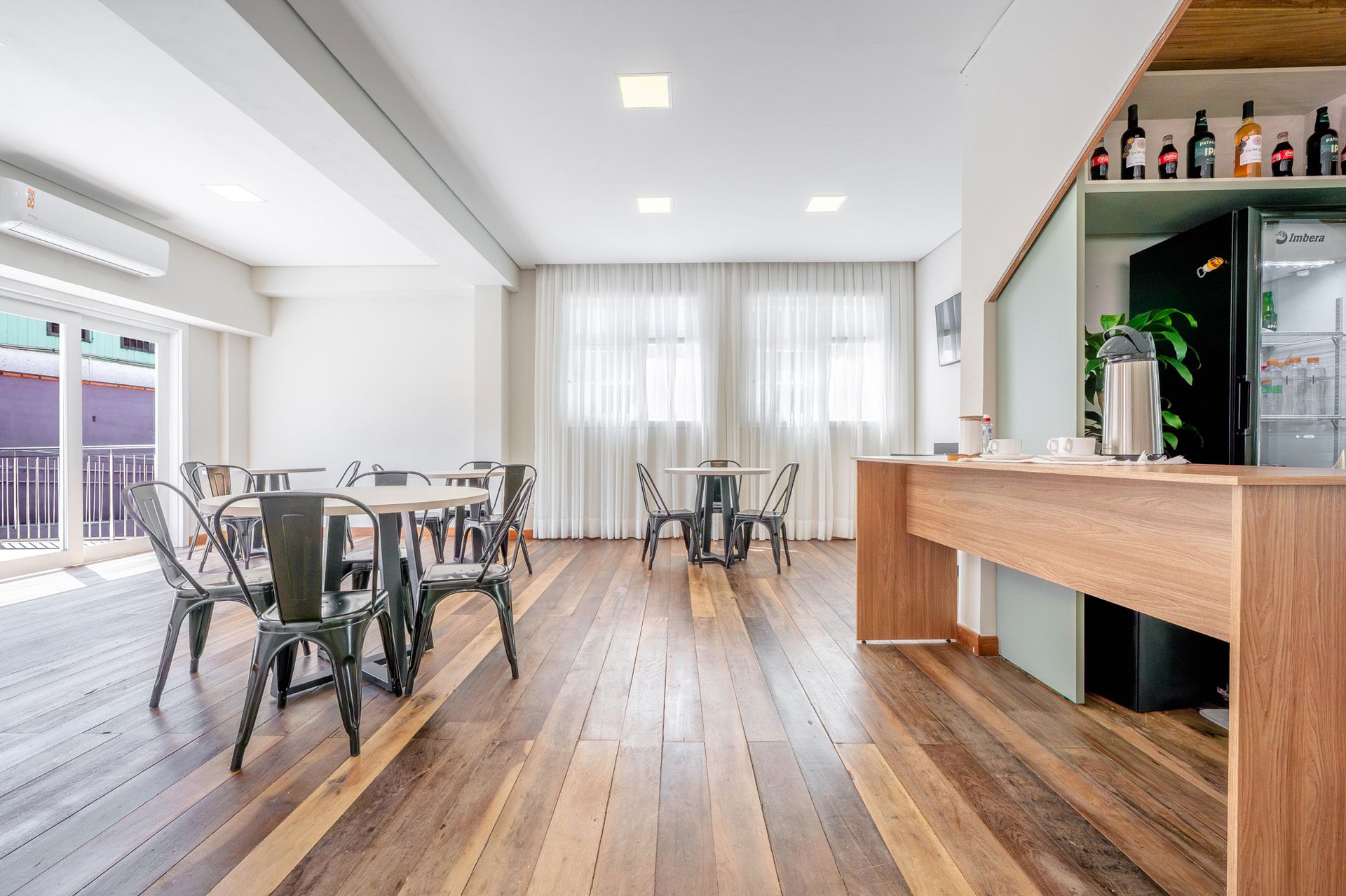 Interior de café com piso de madeira, mesas, cadeiras, área de bar e janelas com cortinas.