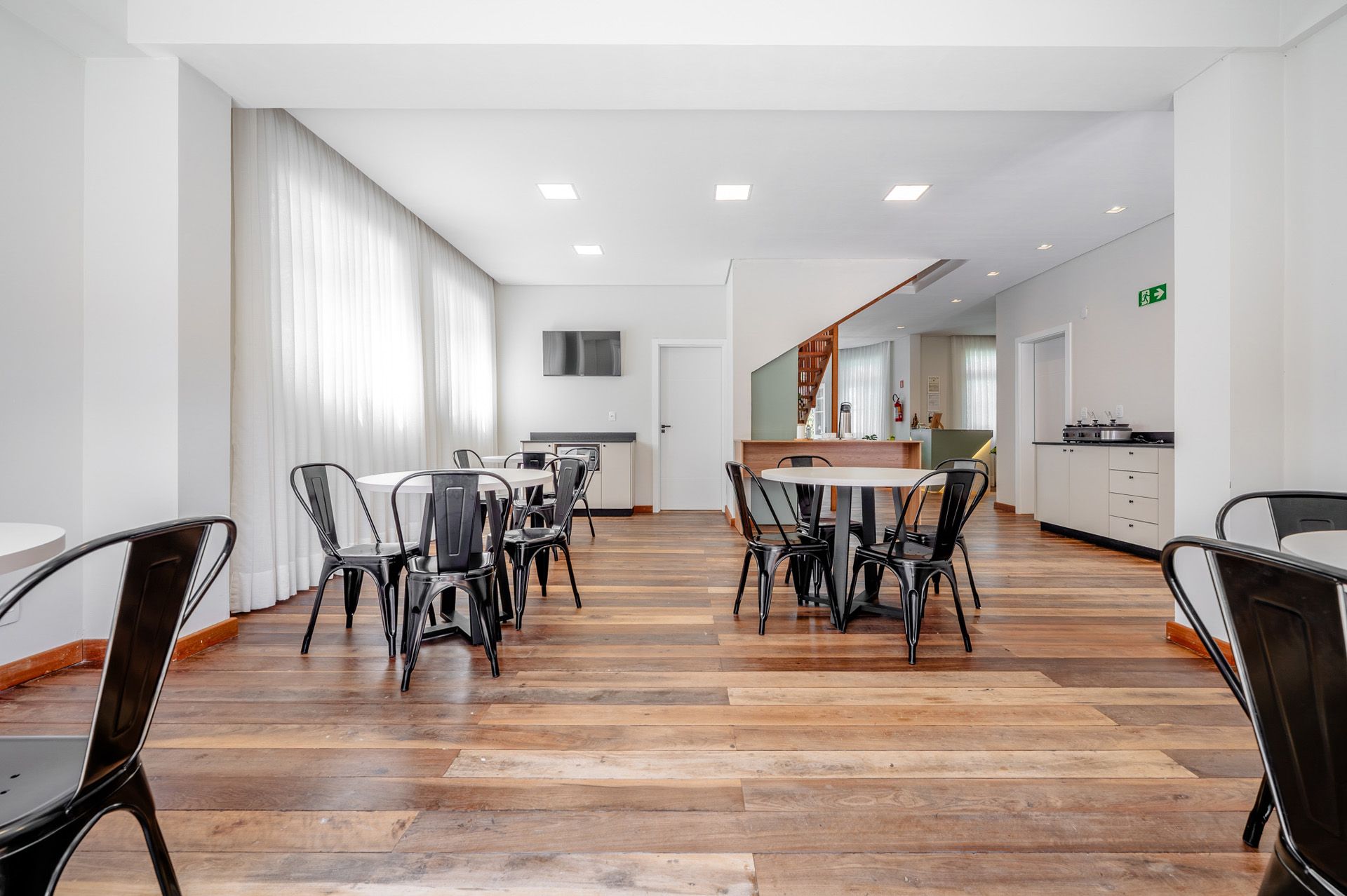 Sala de jantar interna com cadeiras de metal ao redor de mesas sobre piso de madeira, paredes brancas e cortinas.