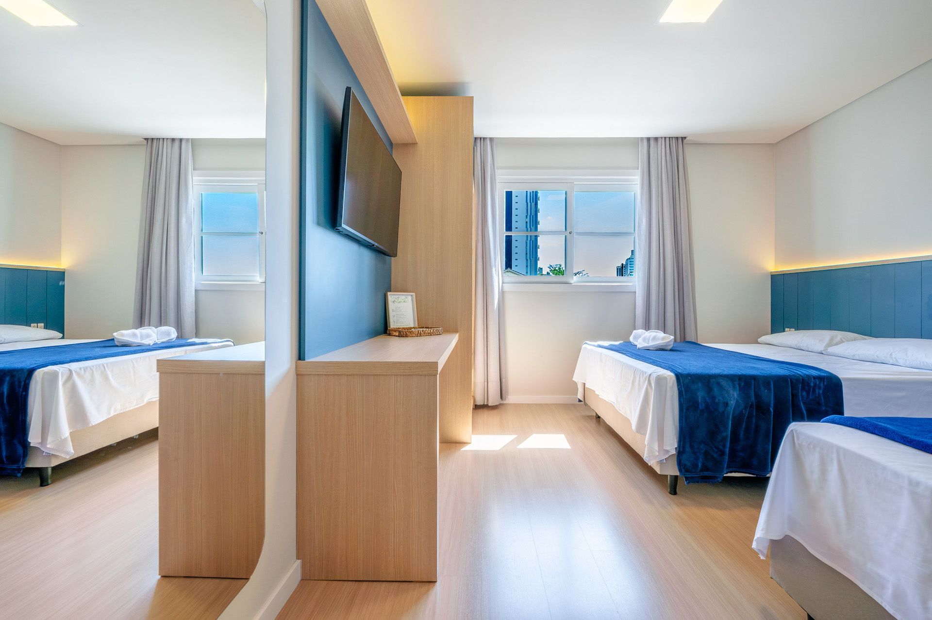 Quarto de hotel com três camas, TV e um grande espelho. O quarto tem piso de madeira clara.