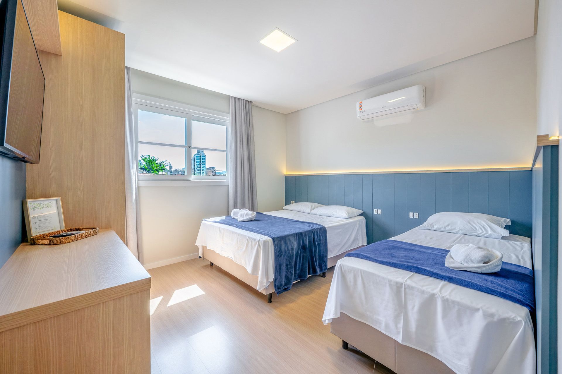 Quarto com duas camas, roupa de cama branca, detalhes em azul, móveis de madeira clara e janela.