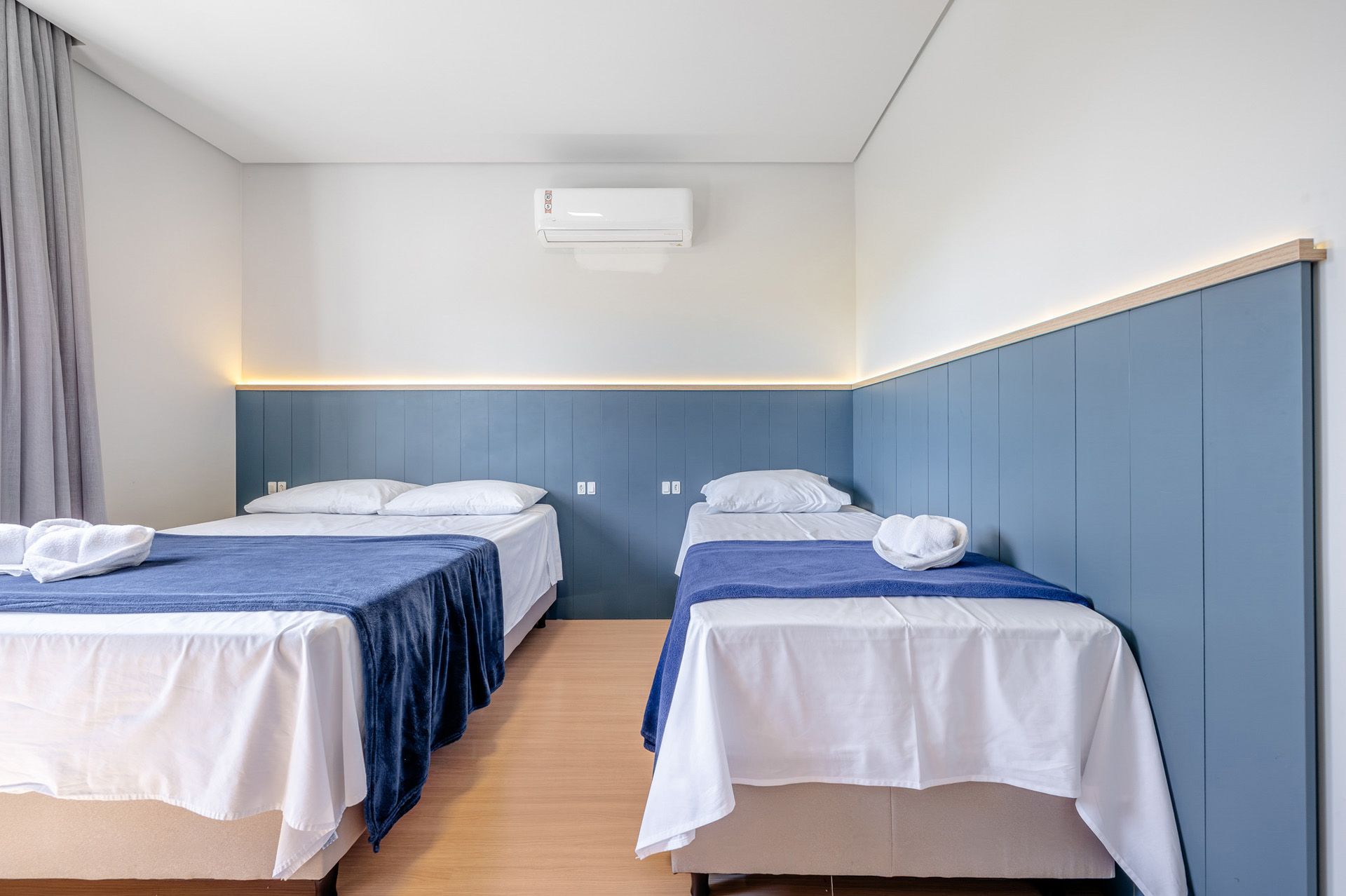 Quarto com duas camas, uma de casal e uma de solteiro; detalhes em azul nas paredes e na roupa de cama; paredes brancas.