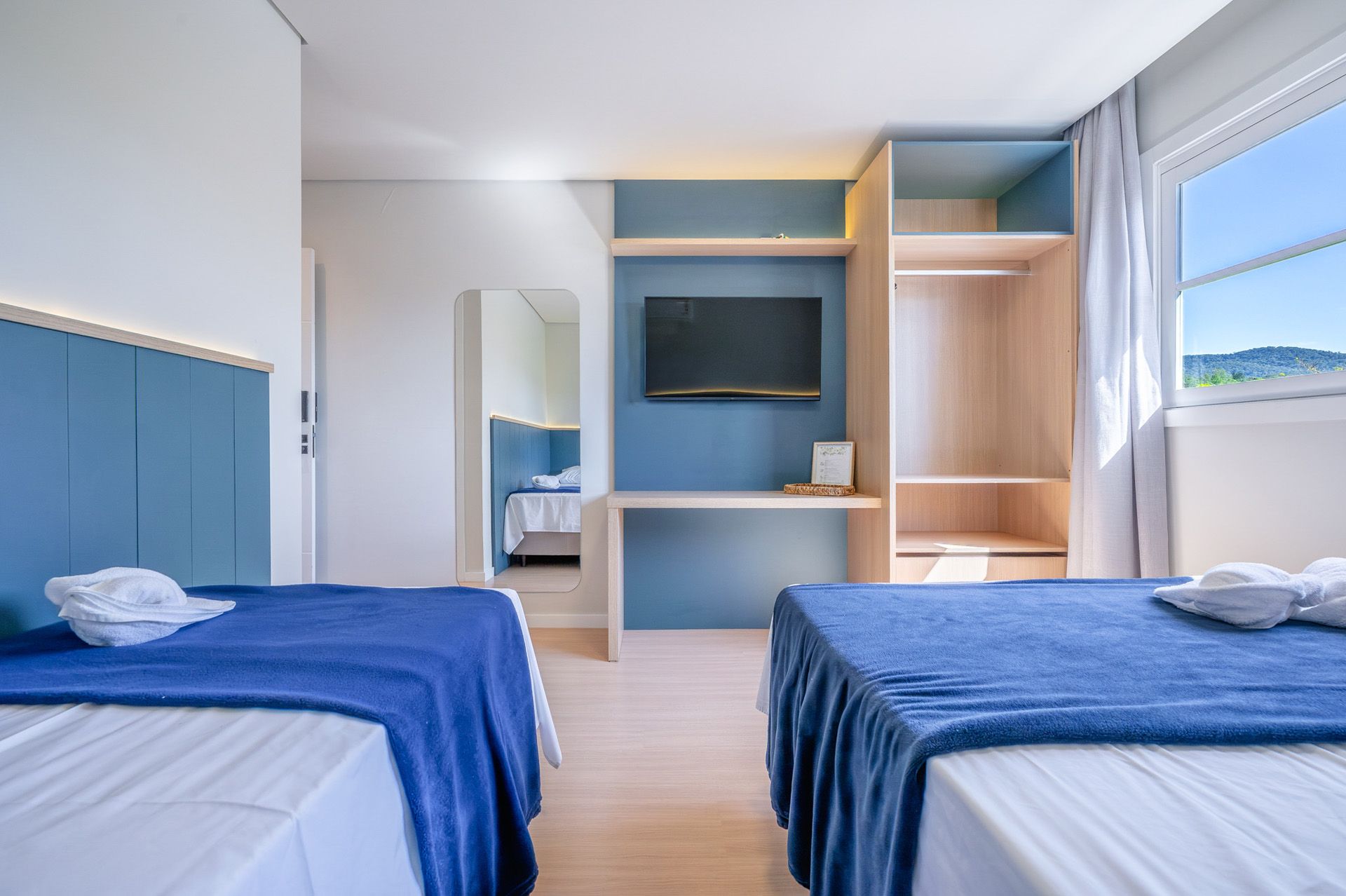 Quarto de hotel com duas camas de solteiro, detalhes em azul, escrivaninha, armário e vista para a janela.