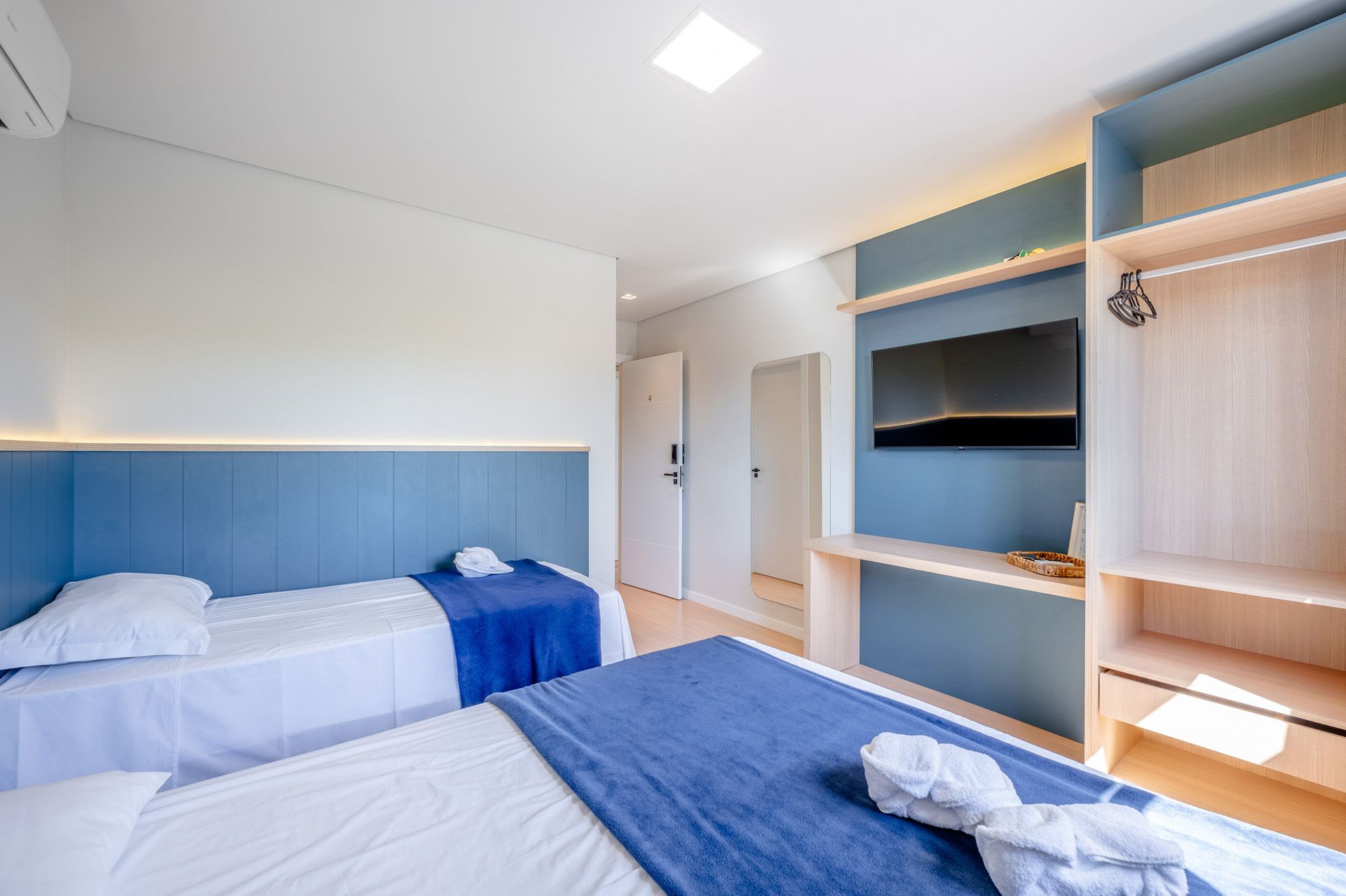 Dois quartos de hotel com duas camas de solteiro, detalhes em azul e uma TV fixada na parede.