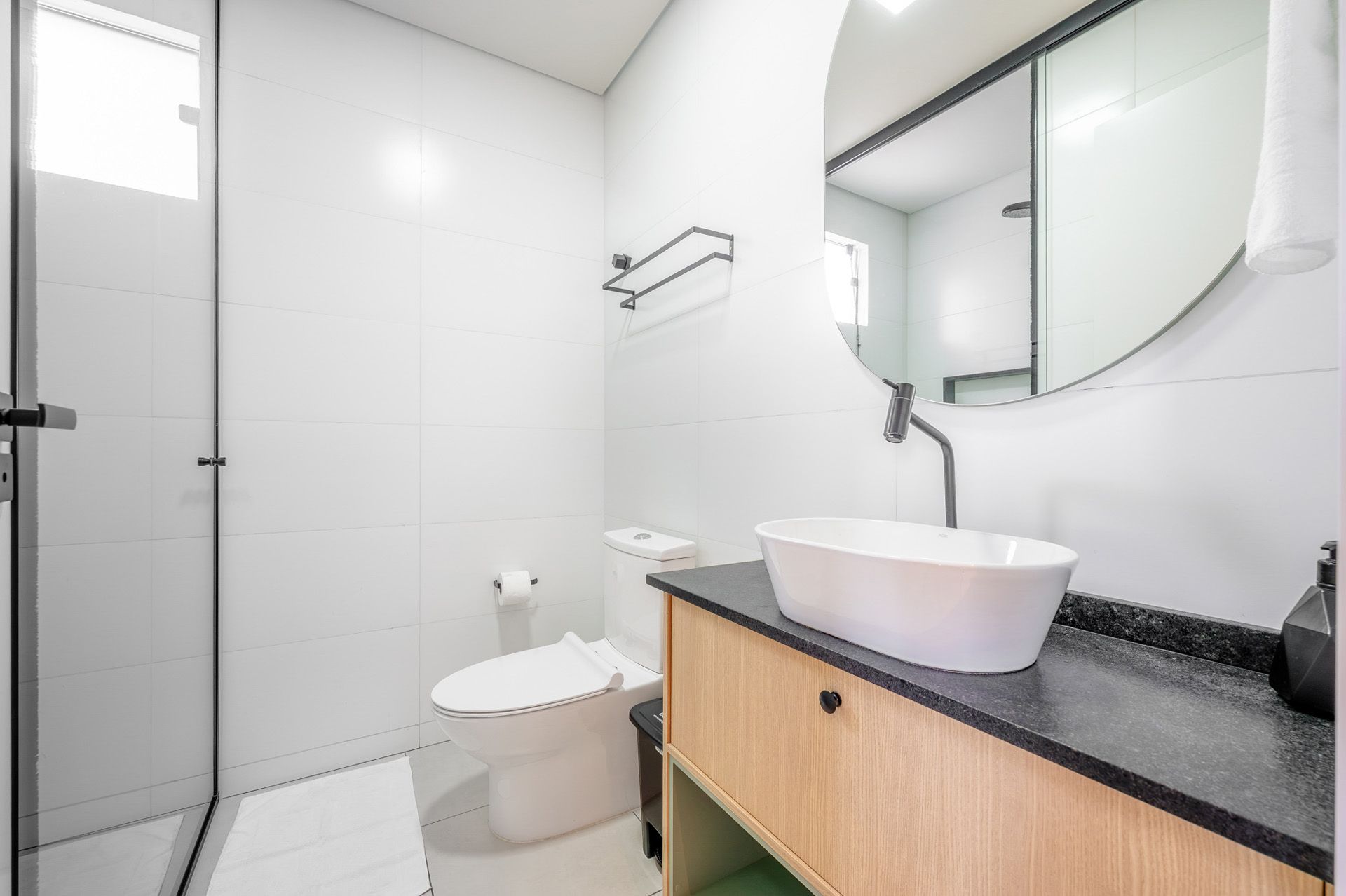 Banheiro branco e iluminado com pia, vaso sanitário e chuveiro. Metais sanitários pretos e bancada de madeira.