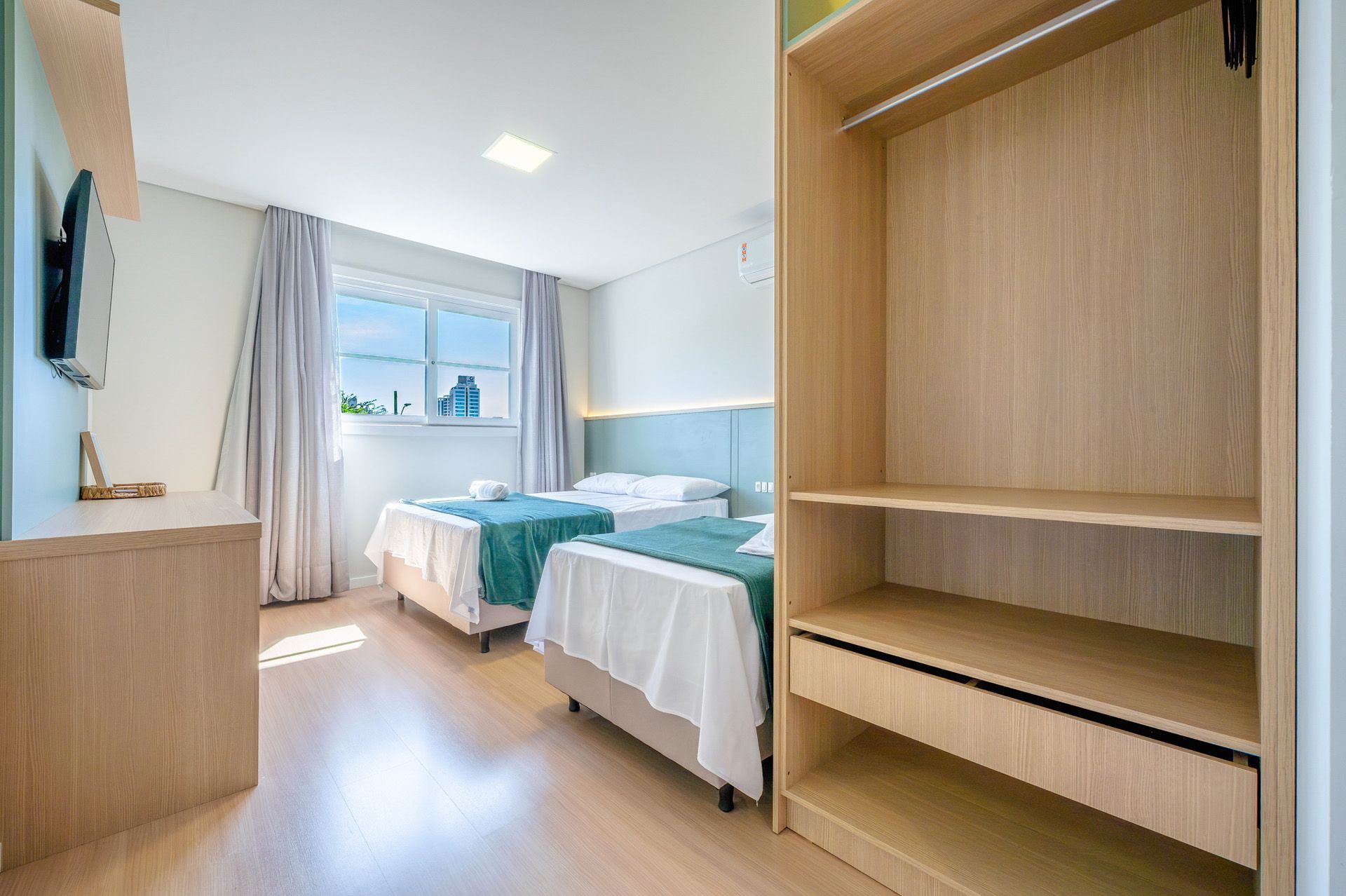 Quarto com duas camas, um guarda-roupa e uma cômoda; paredes e cortinas em tons claros.