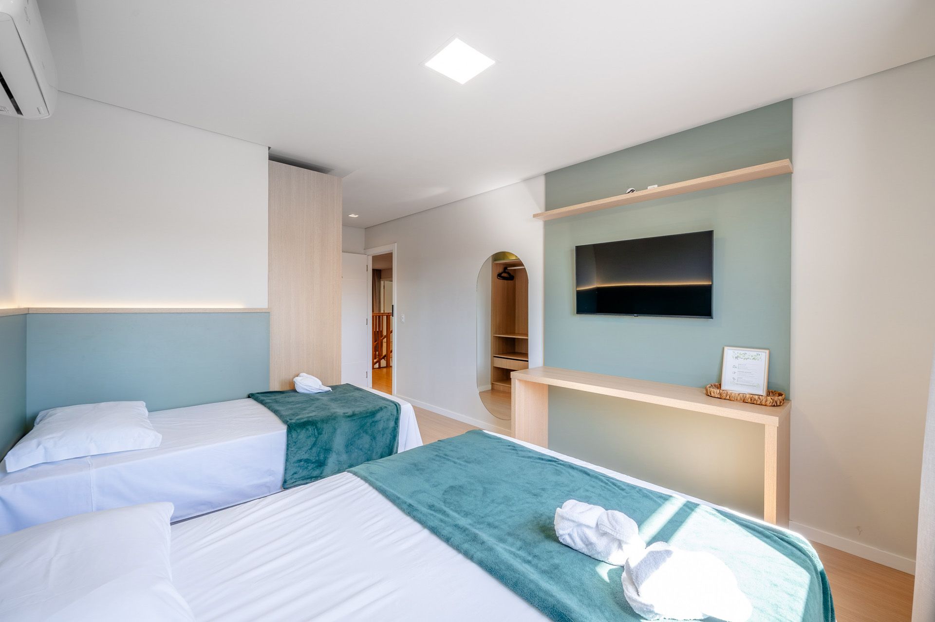 Quarto de hotel moderno com duas camas de solteiro, TV, escrivaninha de madeira clara e detalhes em azul-petróleo.