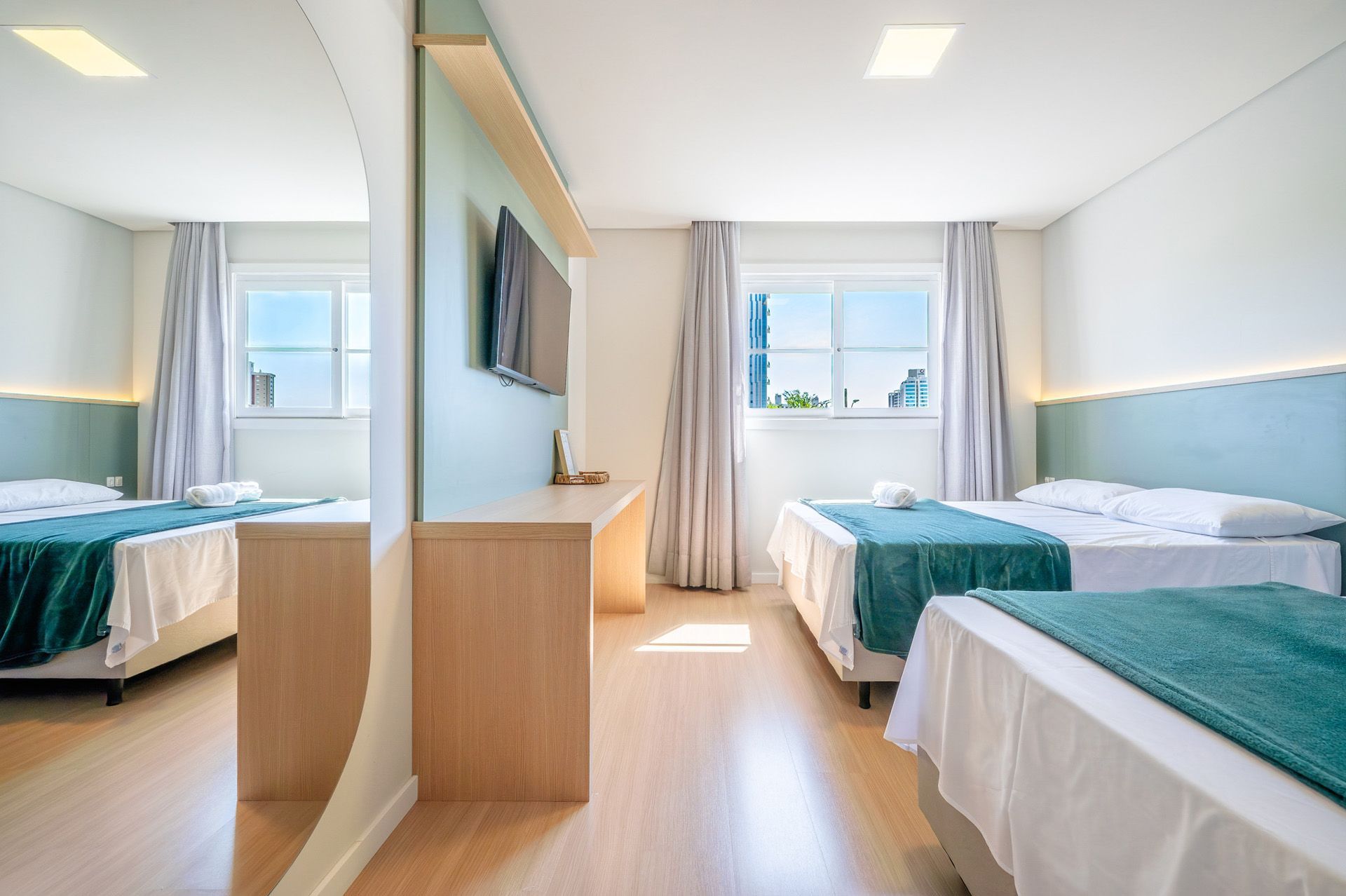 Quarto de hotel com três camas, uma escrivaninha, uma TV e um grande espelho que reflete as camas.