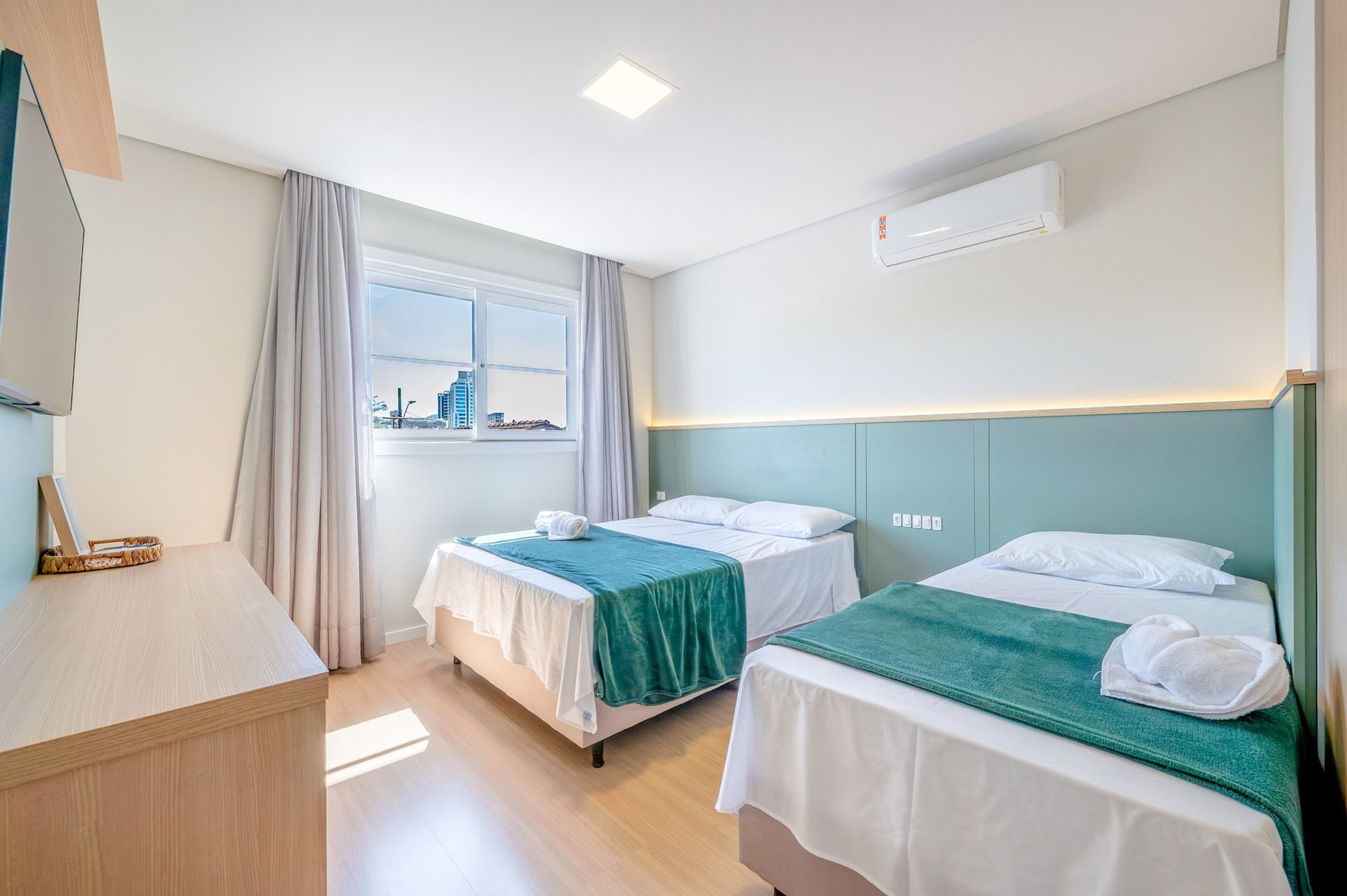 Quarto de hotel com duas camas de solteiro, detalhes em azul-petróleo, janela e móveis de madeira.