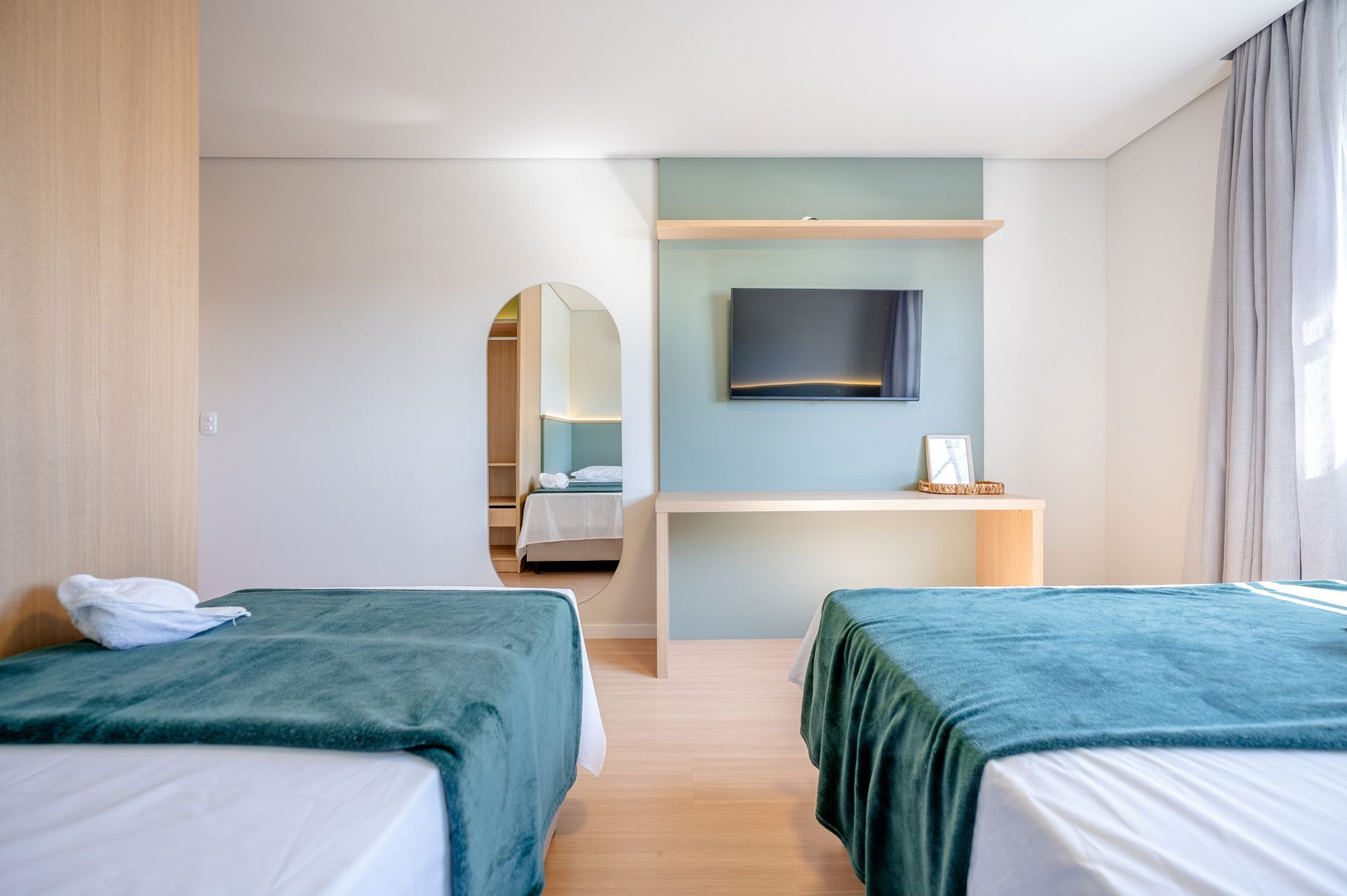 Quarto com duas camas, cobertores verde-azulados, escrivaninha de madeira e TV fixada na parede.