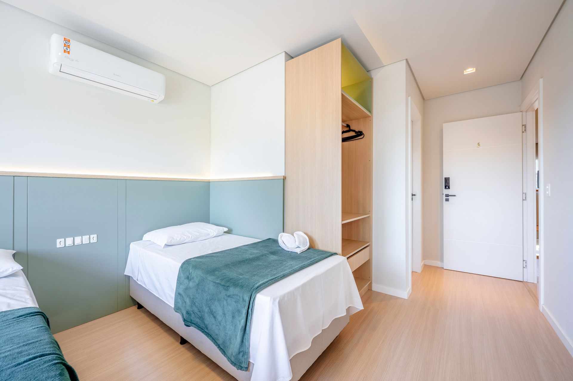 Quarto com cama de solteiro, parede com detalhes em azul-petróleo, armário aberto e porta branca.