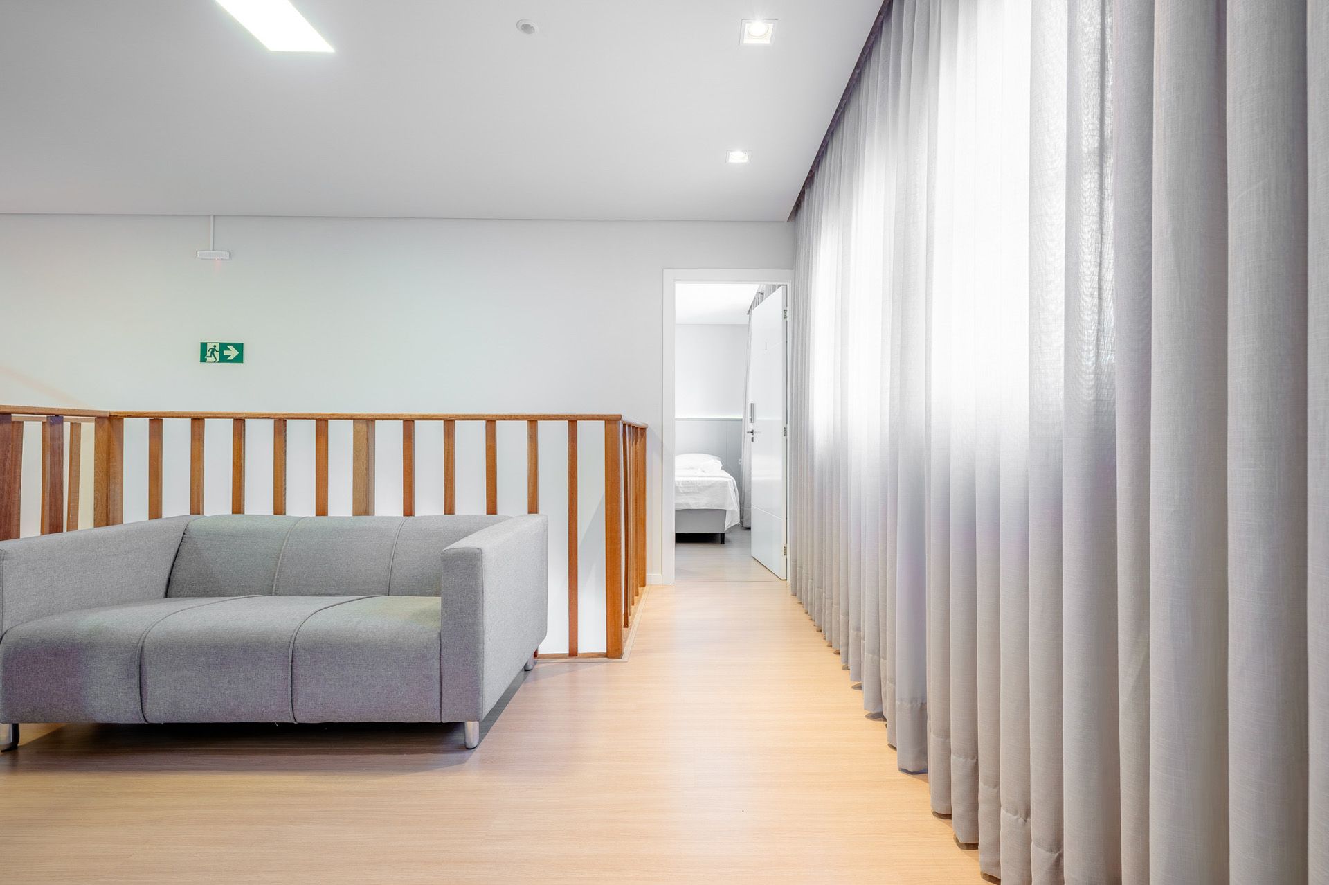 Sofá cinza em uma sala iluminada com piso de madeira clara, um corredor e cortinas translúcidas.