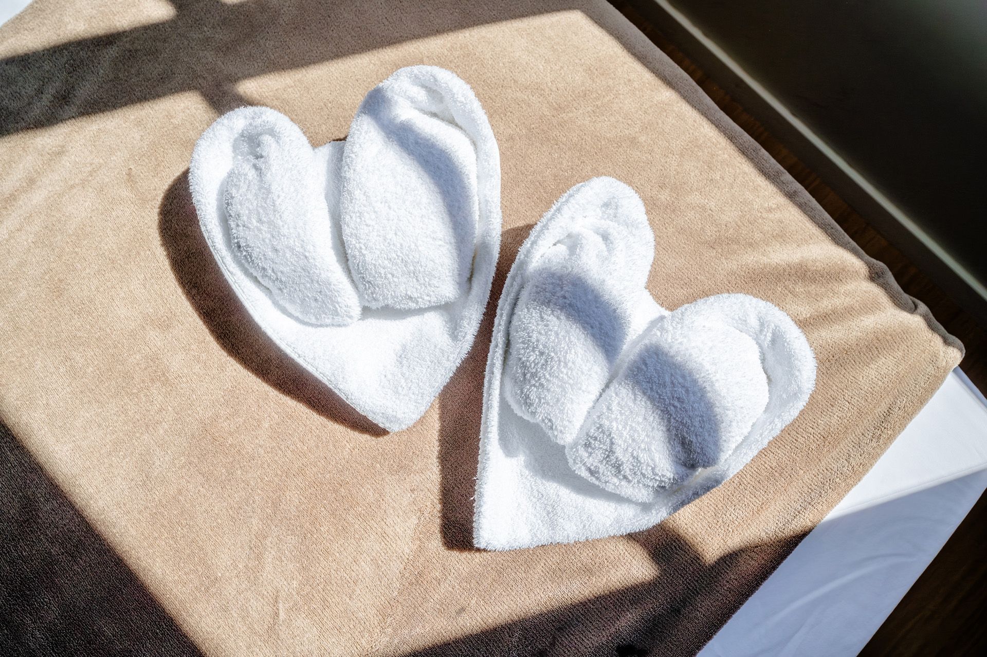 Dois corações de toalha branca sobre uma toalha marrom clara, iluminados pela luz do sol.