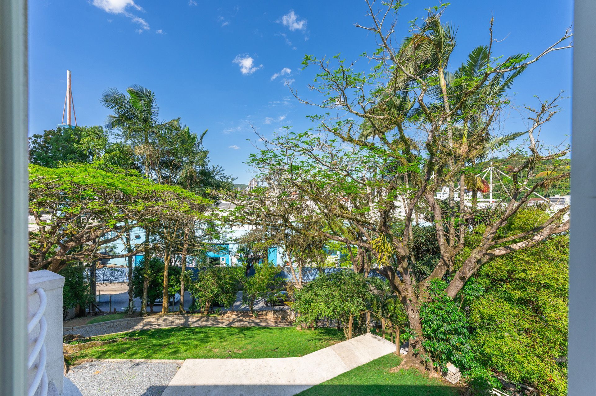 Vista de uma janela com árvores, vegetação e um caminho que leva a edifícios sob um céu azul.