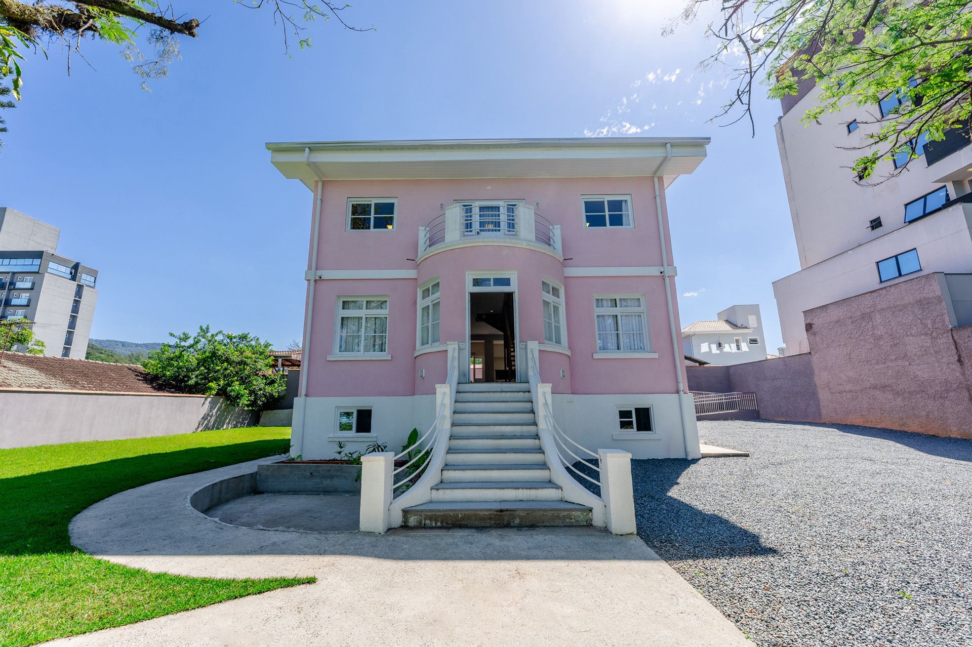 Prédio rosa de dois andares com detalhes em branco, uma escadaria central e uma entrada de cascalho em um dia ensolarado.