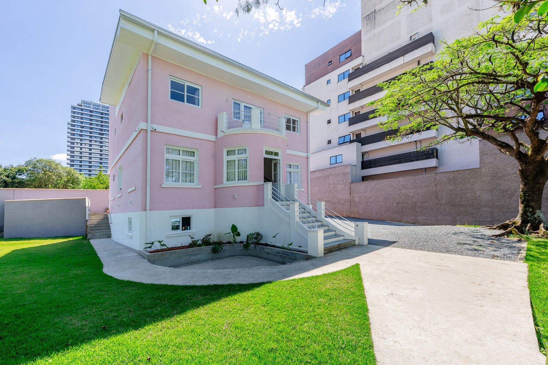 Casa rosa de dois andares com detalhes brancos e gramado verde. Ao lado, um prédio alto e moderno.