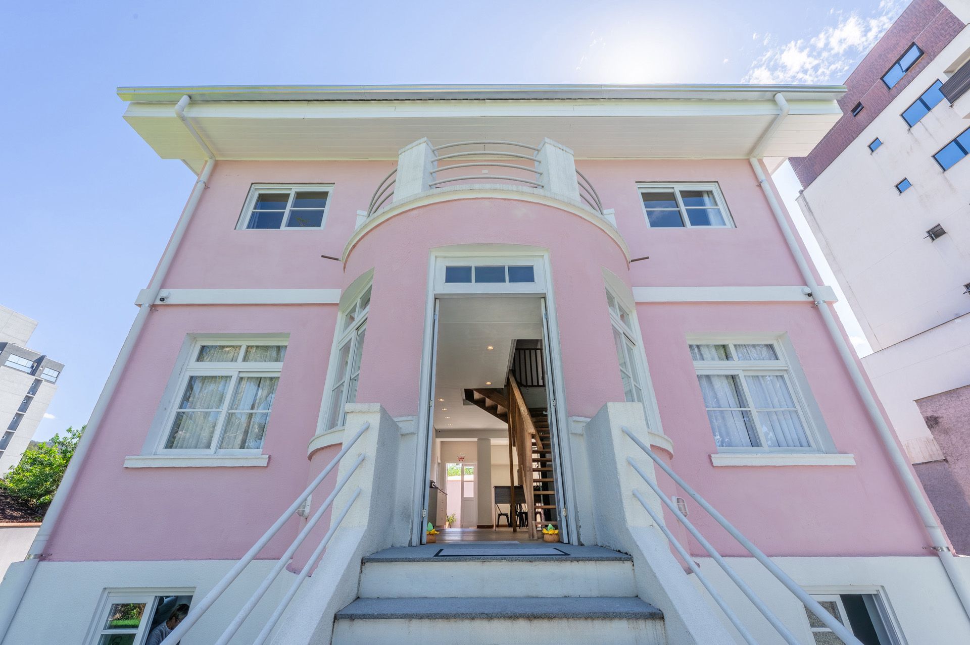 Casa rosa de dois andares com entrada em arco, detalhes em branco e uma pequena varanda. Uma escada leva à porta da frente.