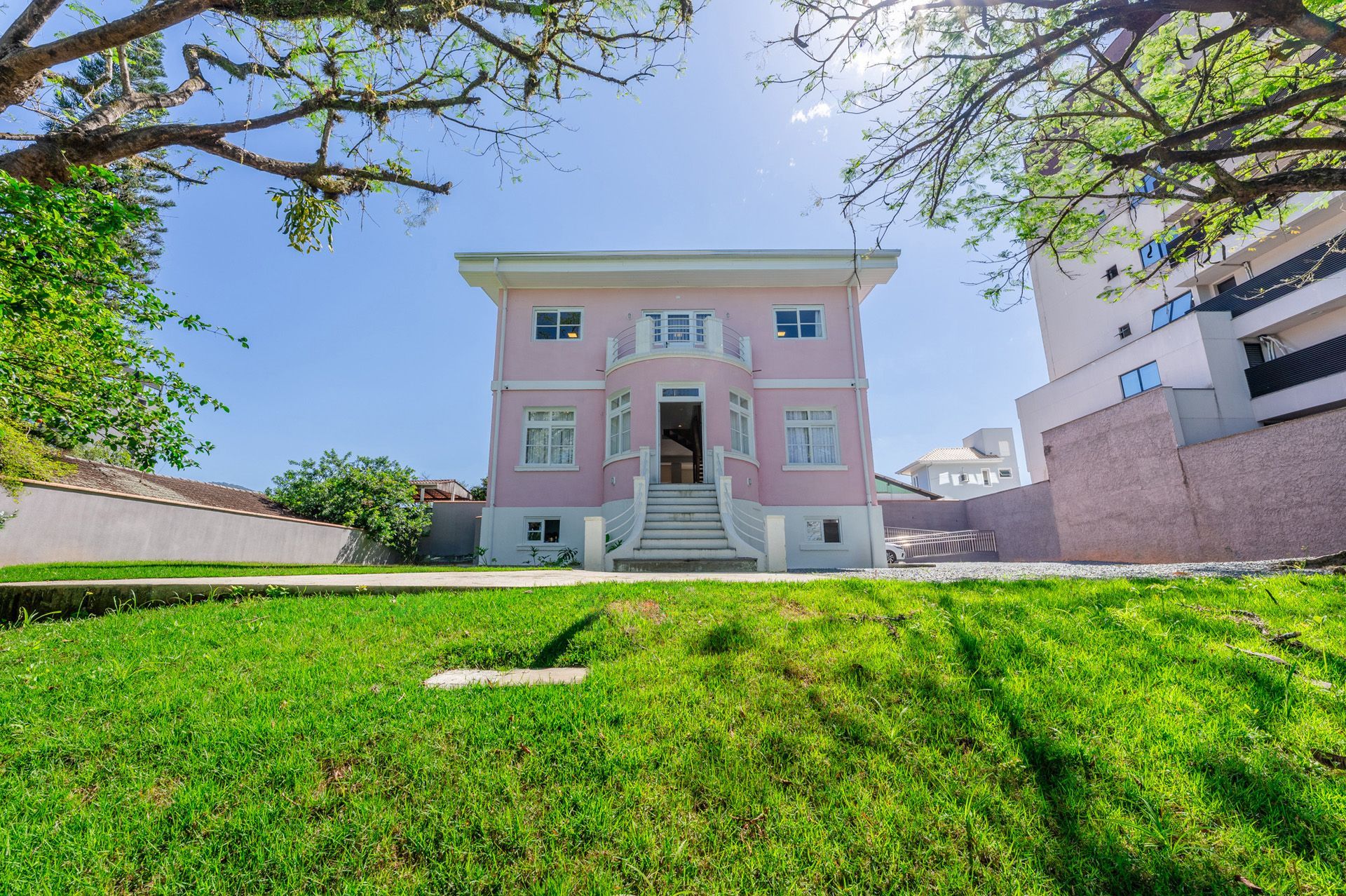 Casa rosa de dois andares com varanda e degraus brancos, situada em um gramado verde com galhos de árvores por cima.