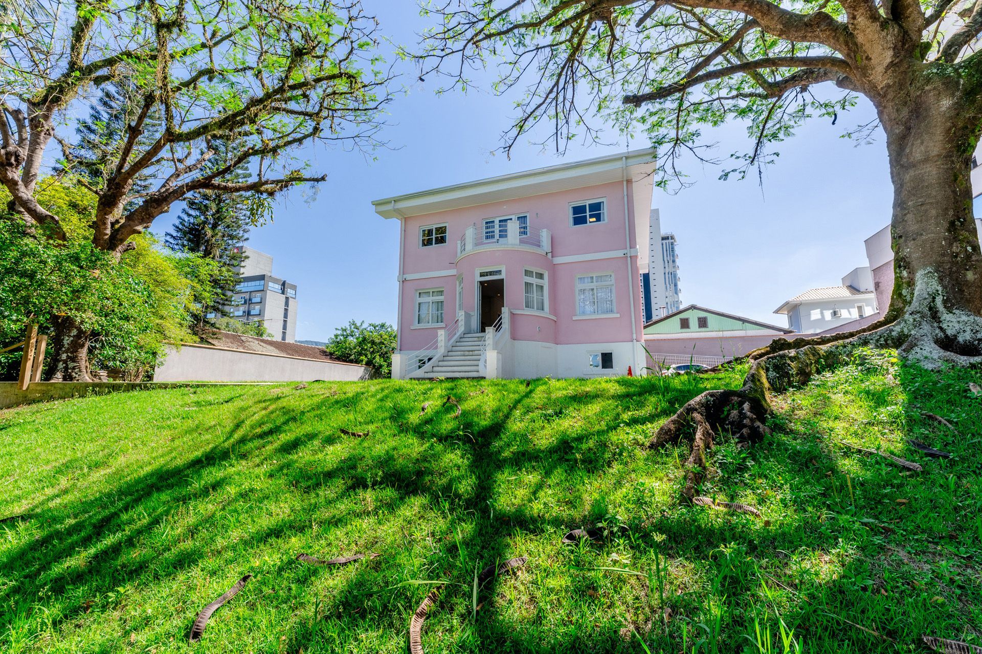 Casa rosa de dois andares em uma colina gramada, emoldurada por árvores. Dia ensolarado.