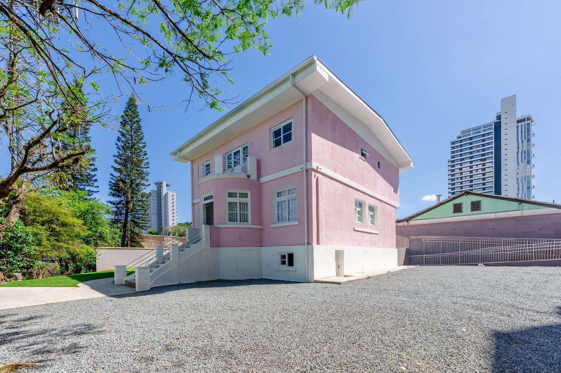 Casa rosa de dois andares com detalhes em branco. Entrada de cascalho, árvores verdes e prédios ao fundo.