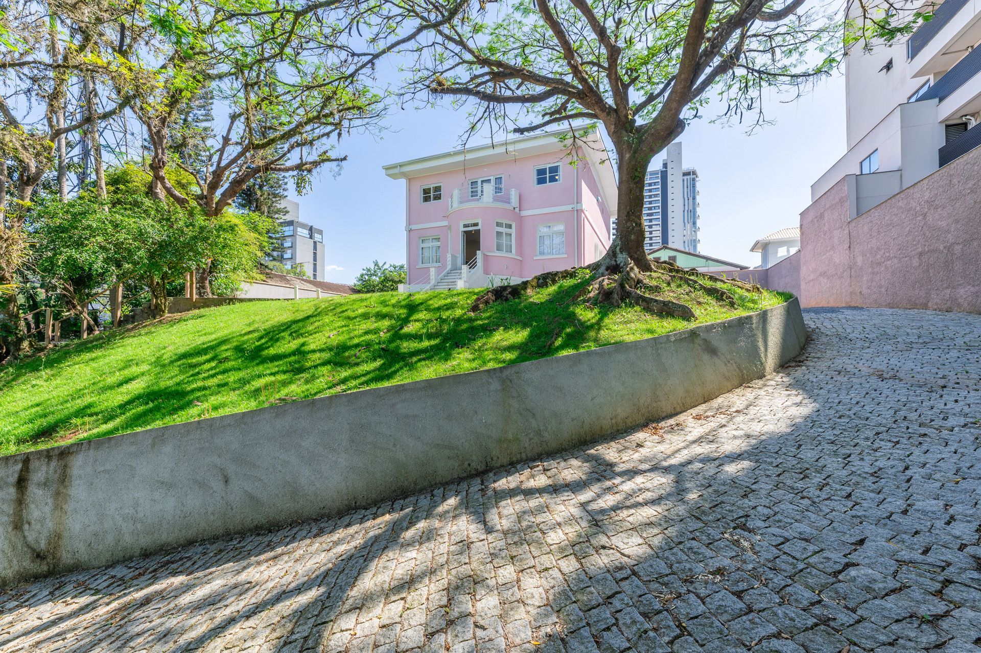 Casa rosa no alto de uma colina, com entrada de paralelepípedos, cercada por grama verde e árvores.