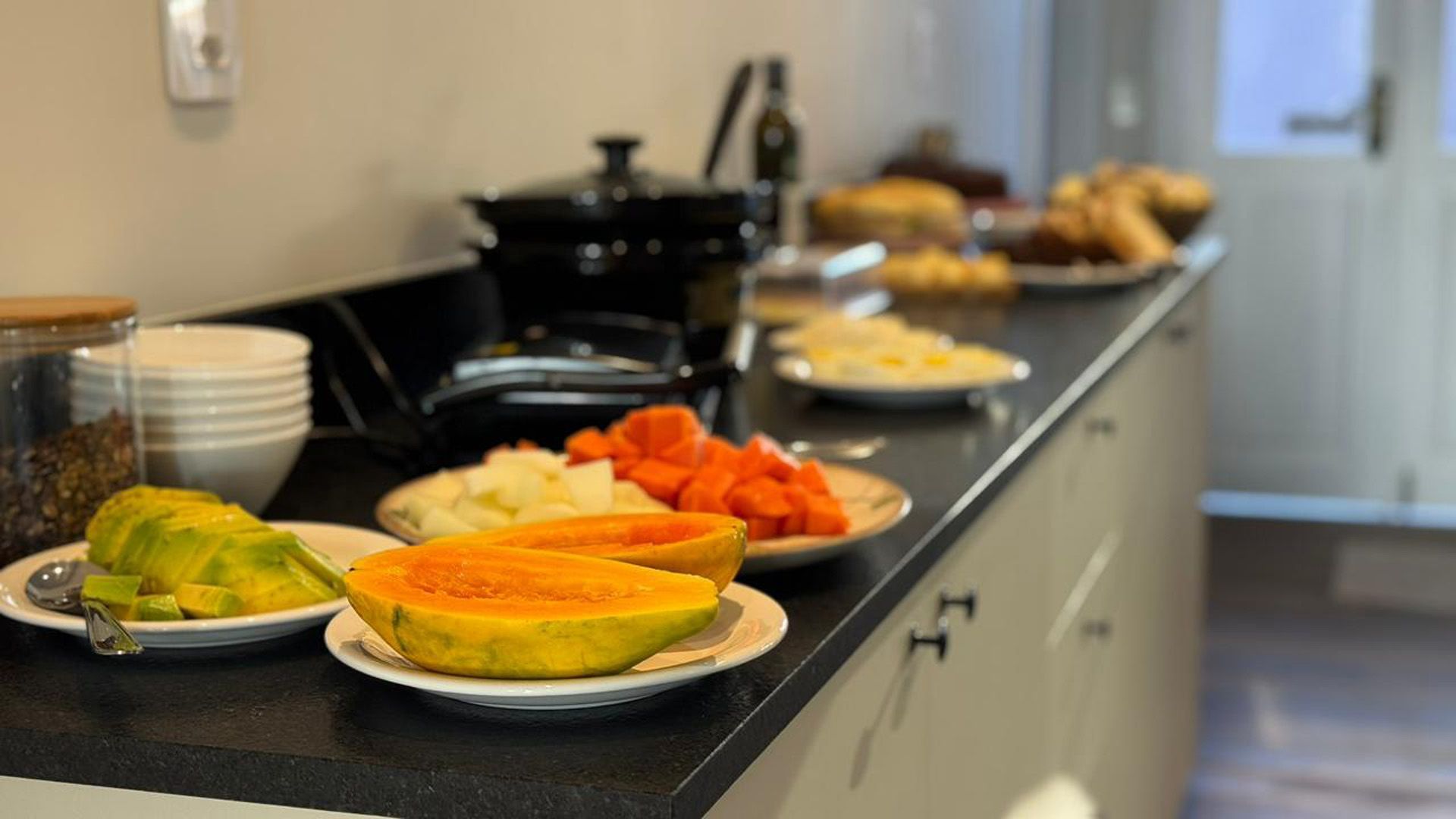Buffet de café da manhã: mamão fatiado, queijo, doces e uma panela elétrica de cozimento lento sobre uma bancada preta.