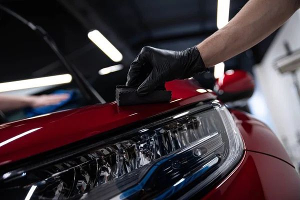 A gloved hand applies a ceramic coating to the red hood of a car, close-up view of the car's headlight.