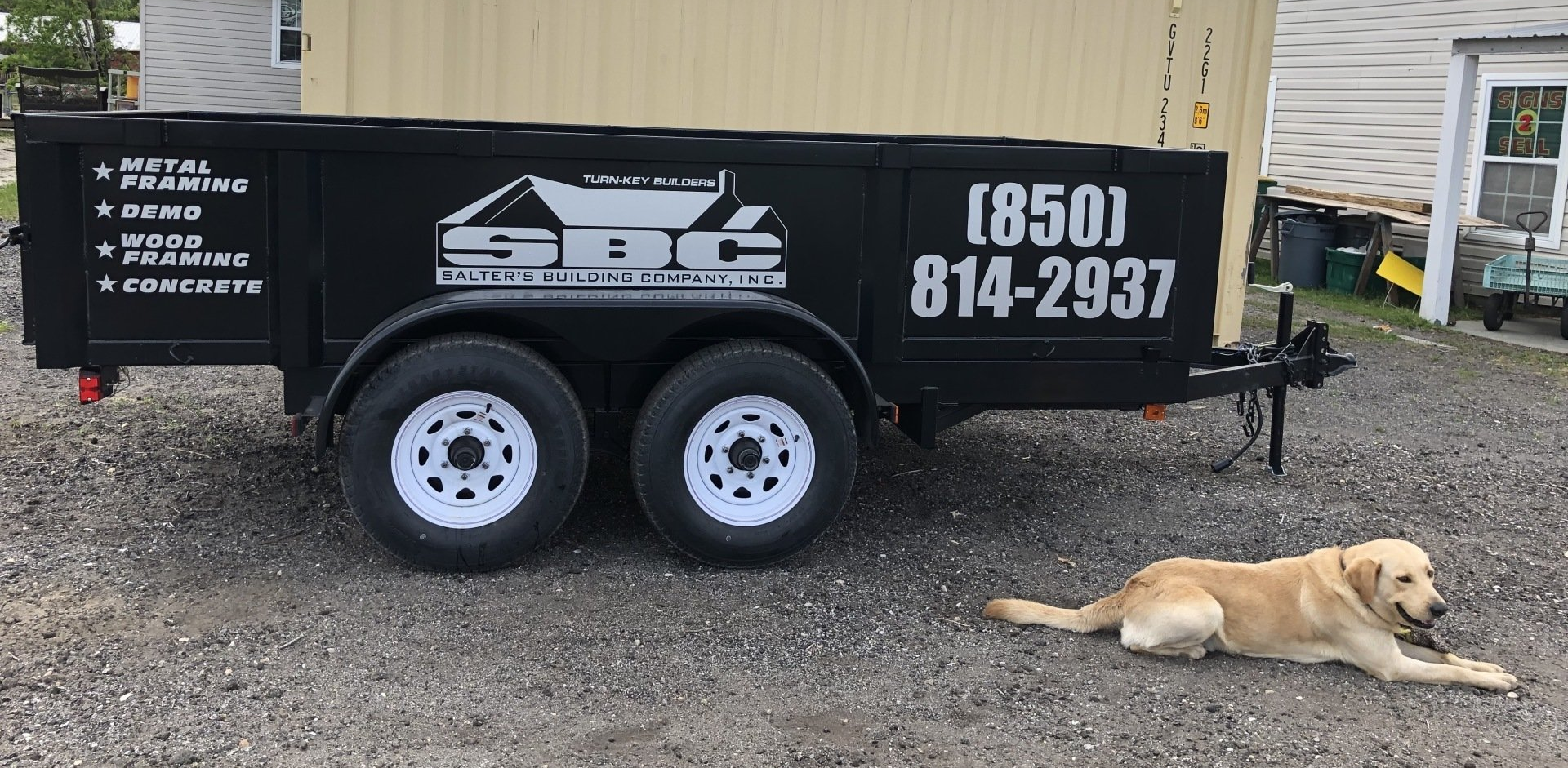 A dog is laying next to a trailer that says sbc on it.