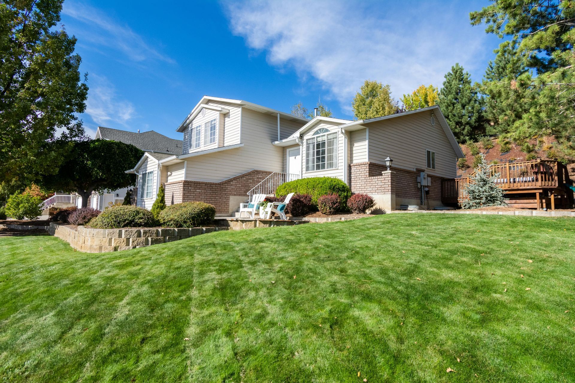 A beige two-story suburban house with a brown brick lower level, a large green lawn, and a wooden deck in the backyard.