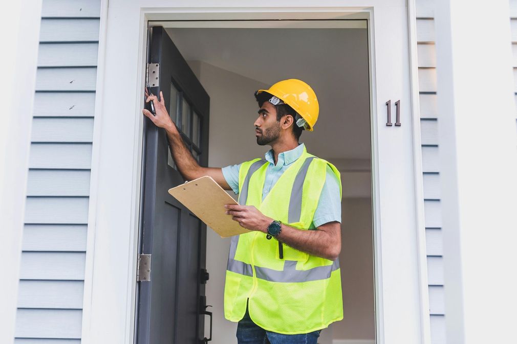 A man wearing a hard hat and safety vest is standing in front of a door holding a clipboard.