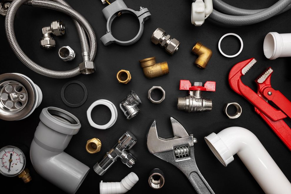 Assorted plumbing tools and pipe fittings on a dark background, including wrenches, valves, and PVC elbows