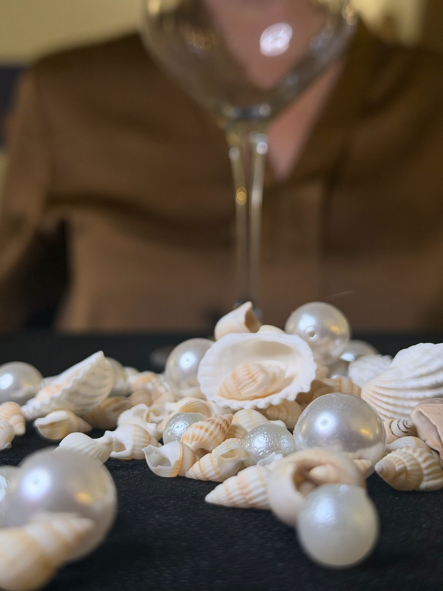 sul tavolo nero vediamo delle perle e conchiglie sparpagliate, un calice vuoto e una donna di sfondo