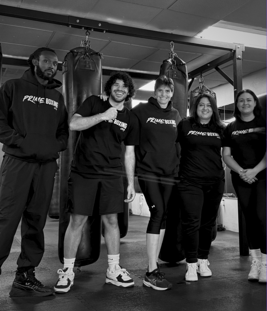 Five people in black athletic wear pose in a boxing gym. Two men and three women smile in front of punching bags.