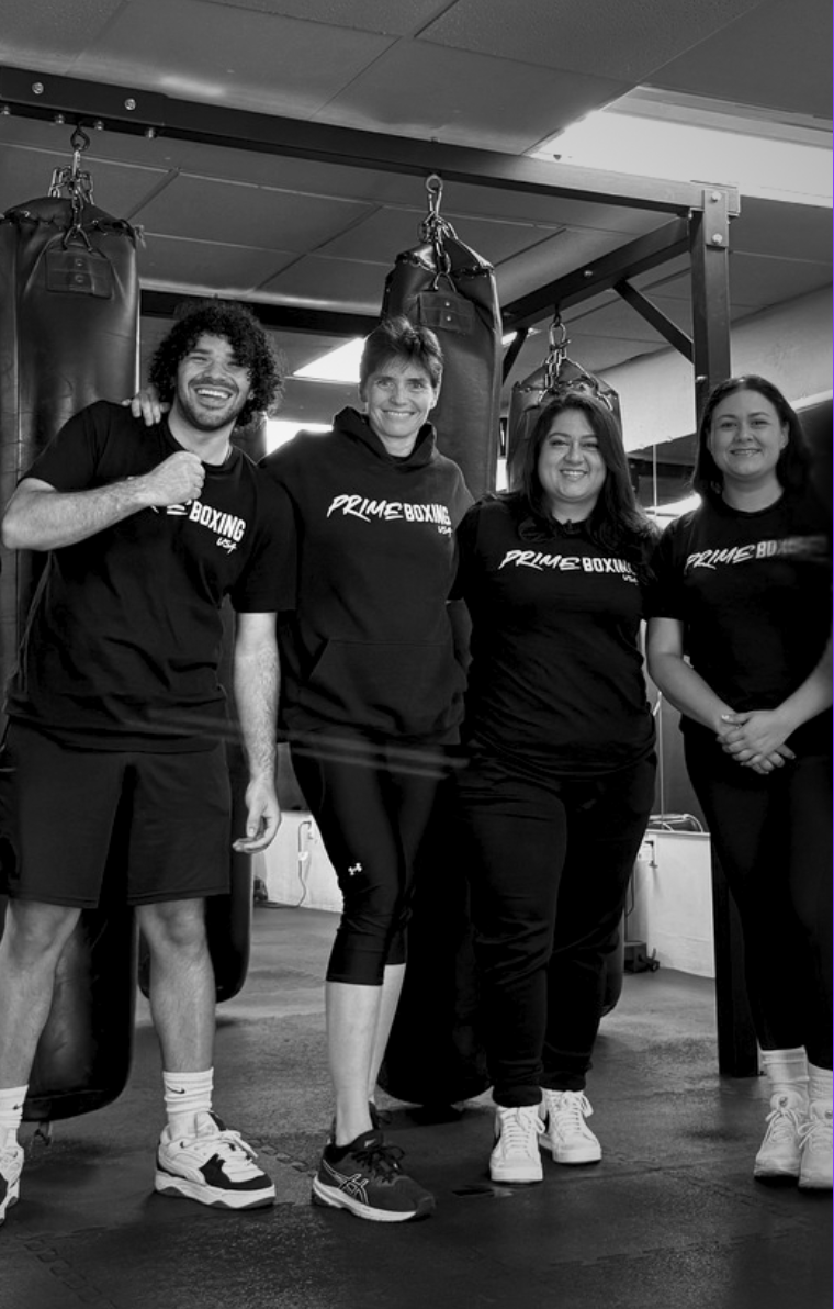 Five people in black athletic wear pose in a boxing gym. Two men and three women smile in front of punching bags.