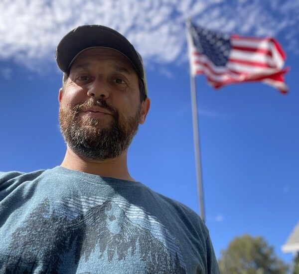A man with a beard is standing in front of an american flag.