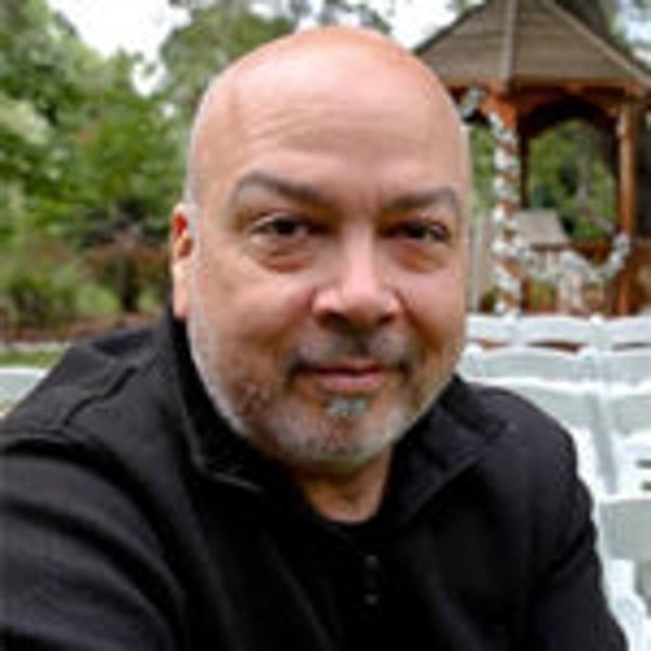 A bald man with a beard is sitting in front of a gazebo.