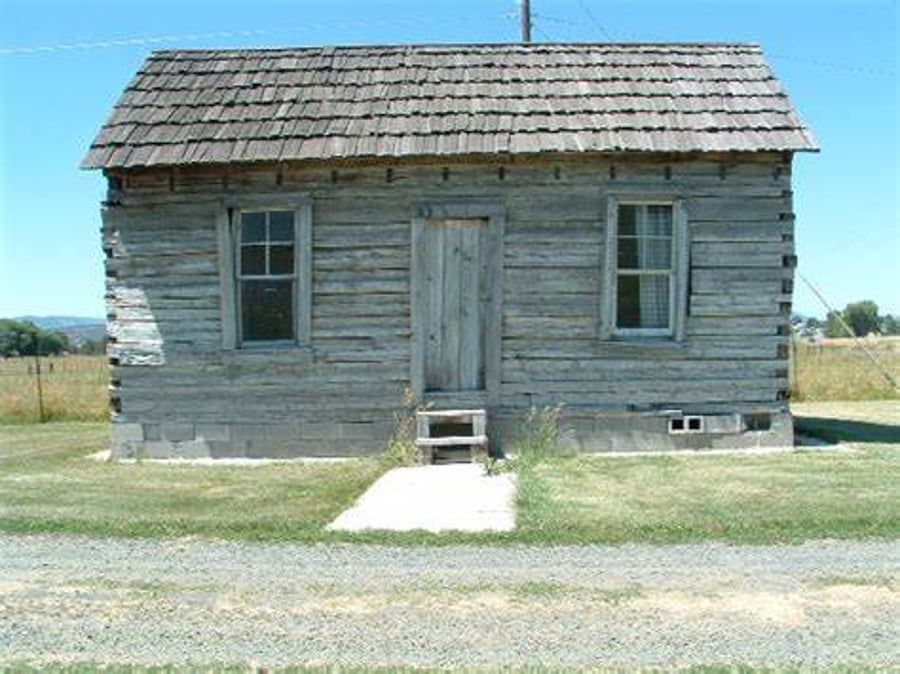 An old log cabin with a shingle roof sits in the middle of a grassy field