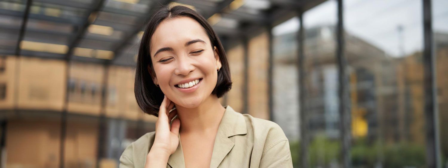 A woman is smiling and touching her face while standing in front of a building.